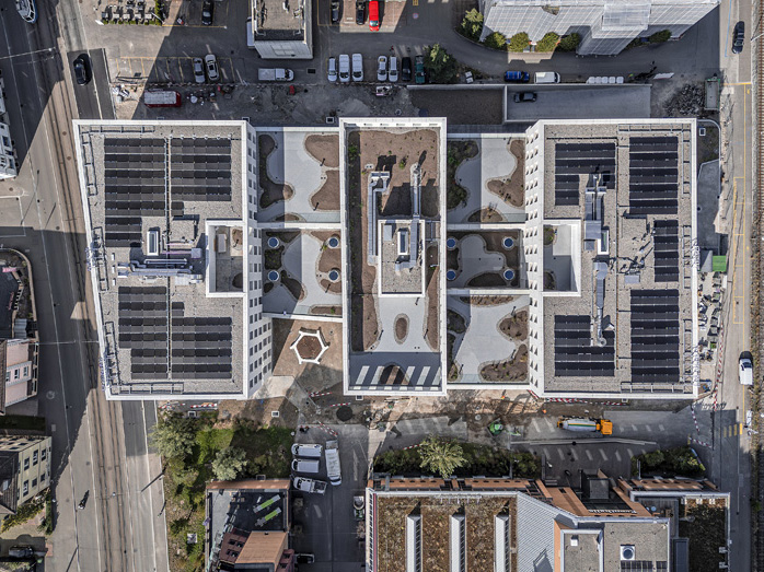 Aerial view of a modern building complex with flat roofs equipped with solar panels and landscaped areas. Surrounding urban infrastructure and streets are visible.