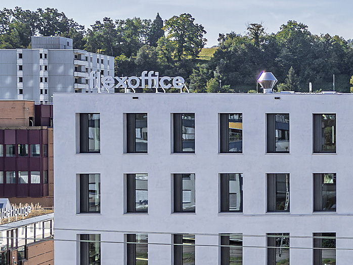 Close-up of a modern office building with a white facade and rectangular windows. The "flexoffice" sign is visible on the rooftop, with greenery and urban buildings in the background.